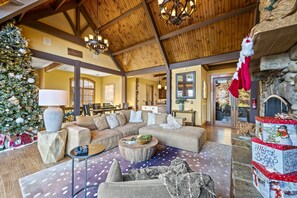 A spacious living room with a large sectional sofa, a round coffee table, and a Christmas tree. There is a fireplace with stockings and a Christmas tree with presents.
