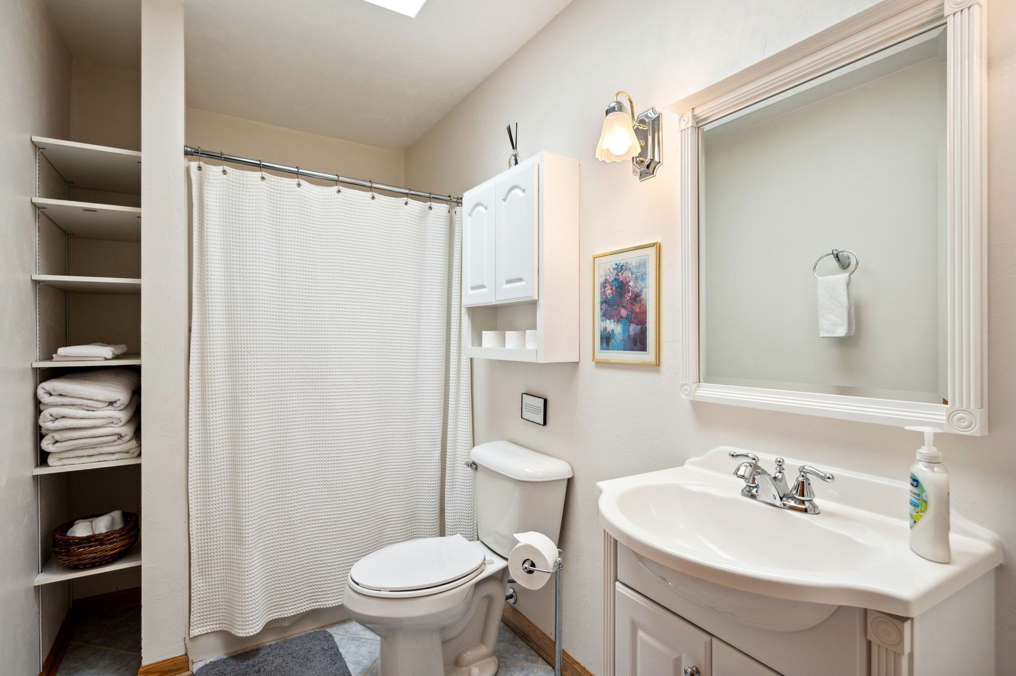 Un baño con inodoro, lavamanos y cortina de ducha. Hay estantes con toallas y un cuadro enmarcado en la pared.