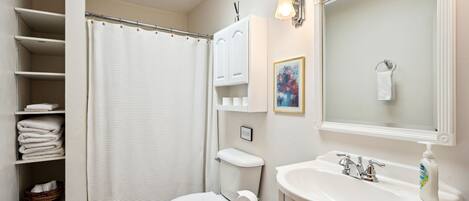Un baño con inodoro, lavamanos y cortina de ducha. Hay estantes con toallas y un cuadro enmarcado en la pared.