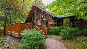 A log cabin with a wooden deck and a brick pathway leading to it. - Charming 3-bedroom Log Cabin by the lake (Egg Harbor)