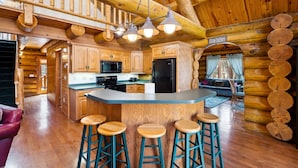 A log cabin interior with a central island kitchen, wooden bar stools, and a cozy seating area. - Charming 3-bedroom Log Cabin by the lake (Egg Harbor)