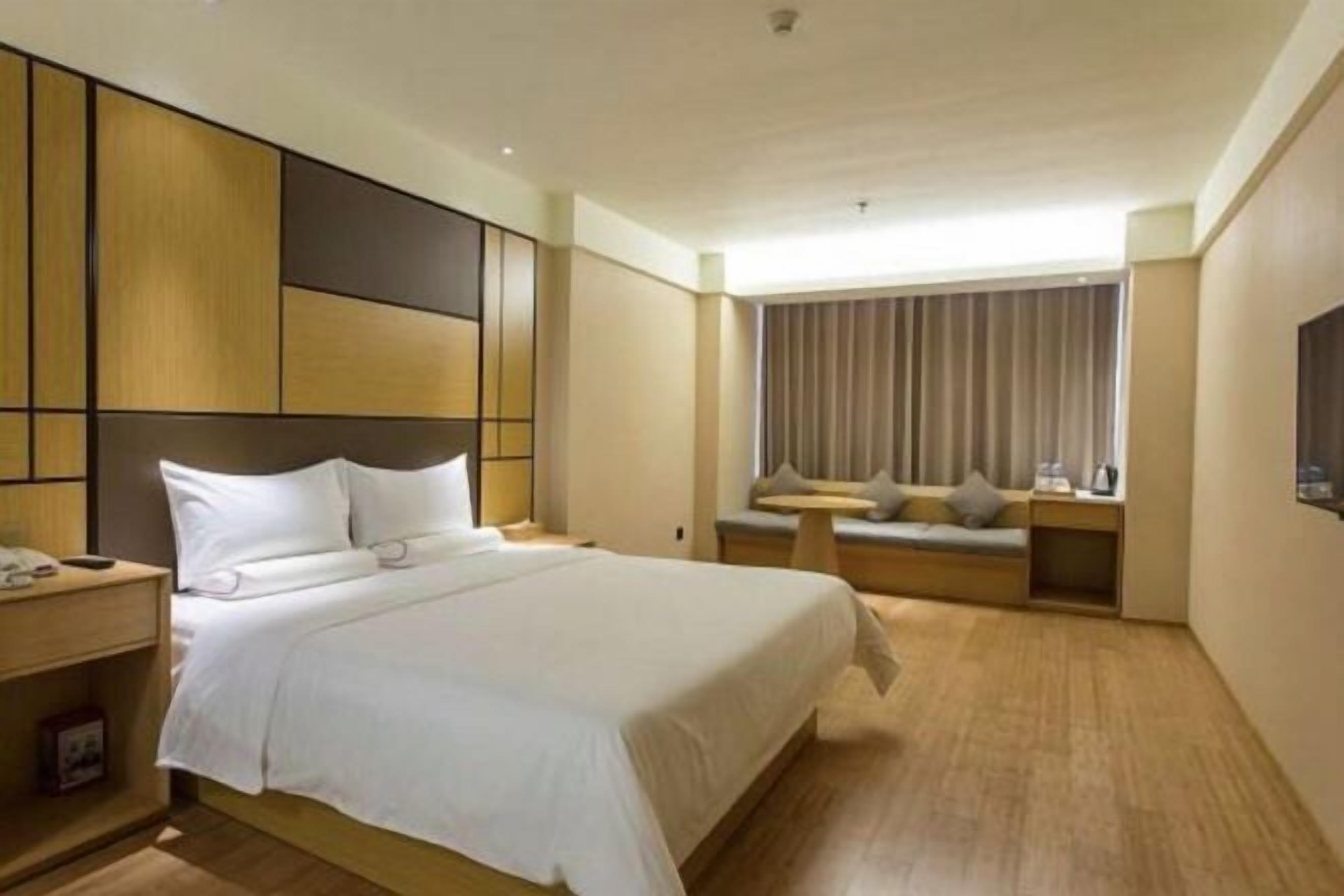 A hotel room with a large bed, a bedside table, a small table, a sofa, and a window with curtains.