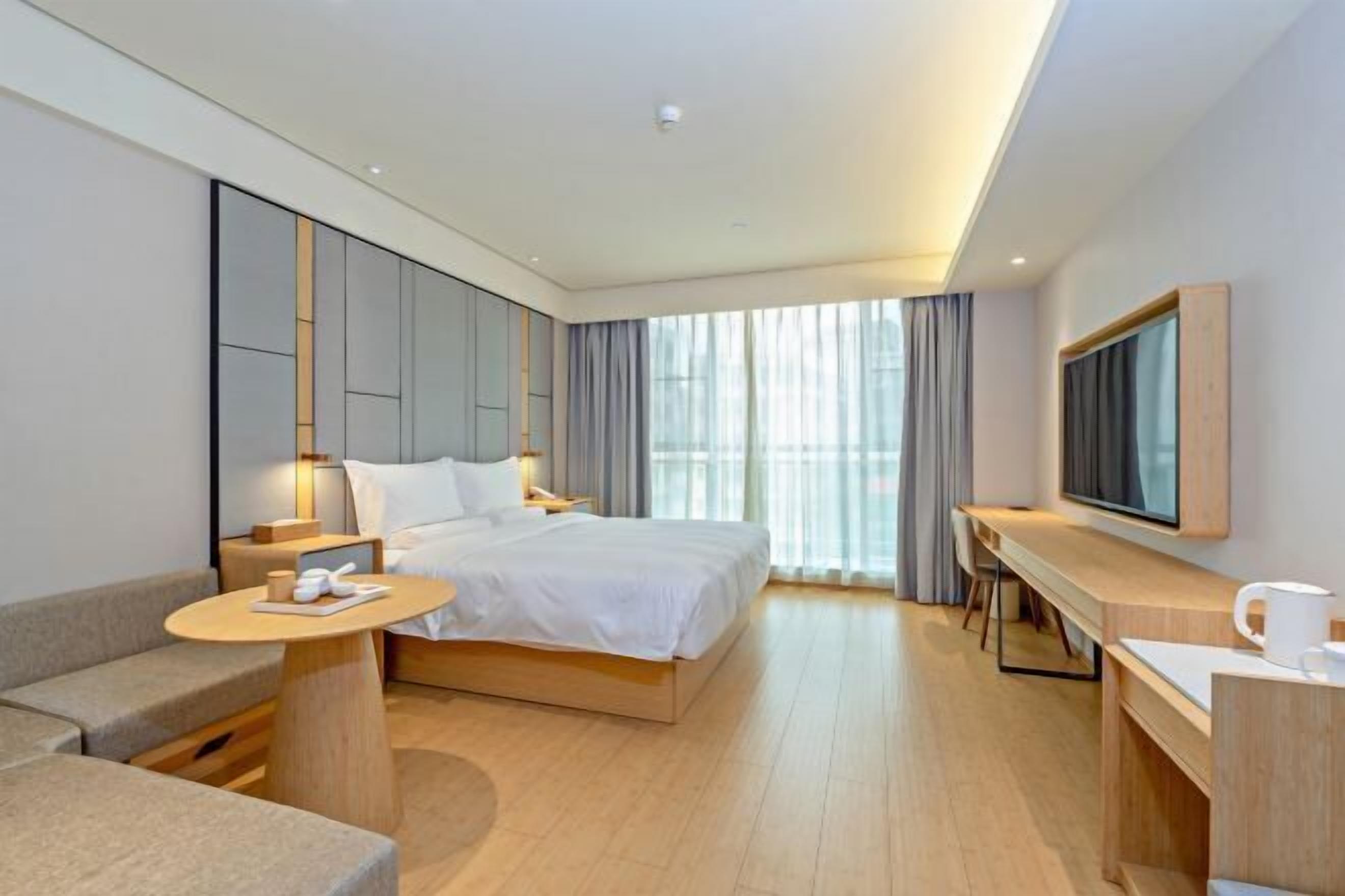A modern hotel room with a large bed, a desk, a TV, and a balcony with curtains.