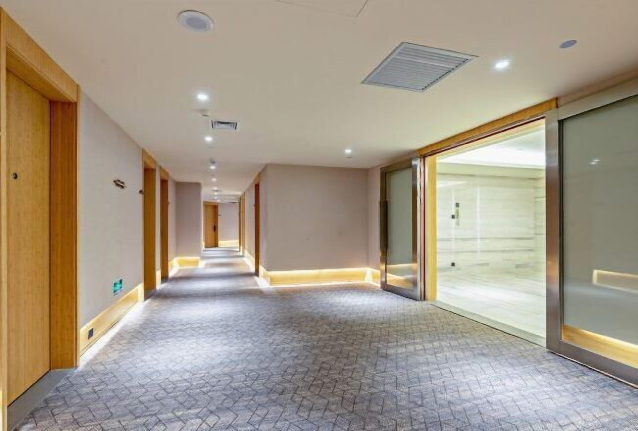 A hotel corridor with a patterned floor, wooden doors, and a glass elevator.