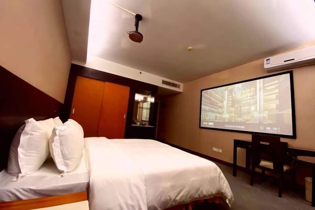 A hotel room with a bed, a desk, a projector screen, and an air conditioning unit.