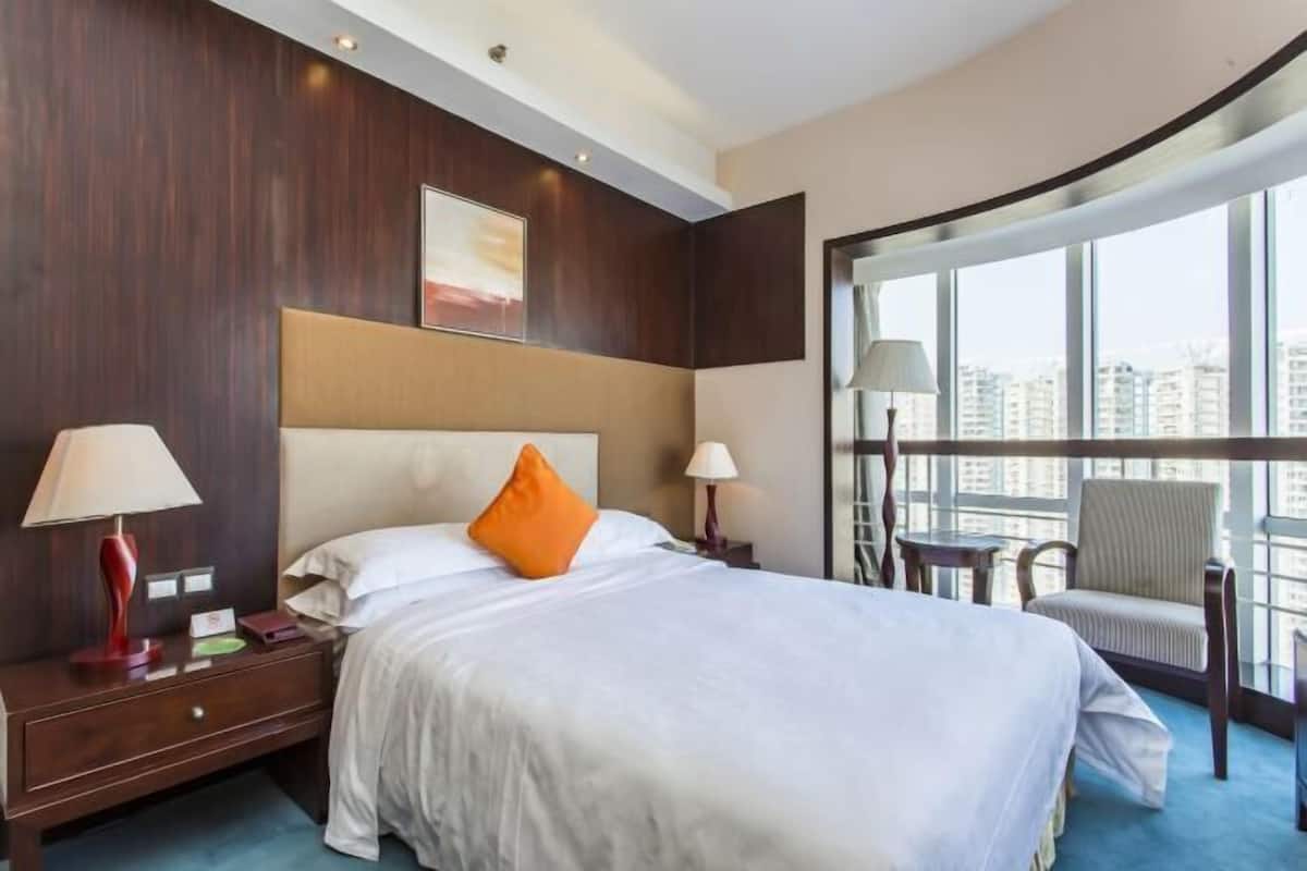 A hotel room with a large bed, a nightstand, a chair, and a view of the city through large windows.