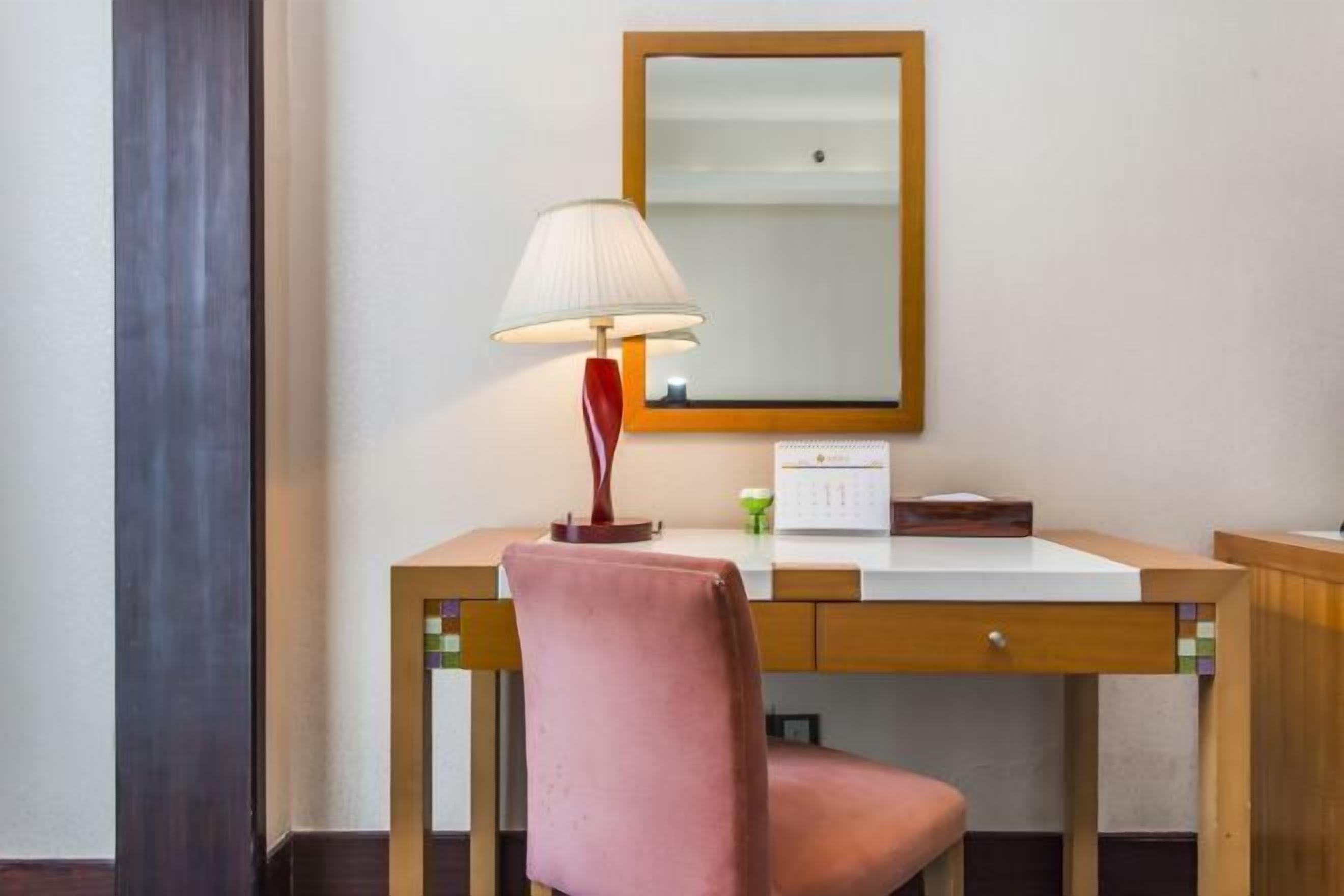 Habitación de hotel con un escritorio de madera, una lámpara, un espejo y una silla rosa.