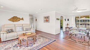 A spacious living room with a beige sofa, a coffee table, and a patterned rug. There is a dining area with a table and chairs, and artwork on the walls. - Beach Views - Spacious home by the ocean (Lennox Head)