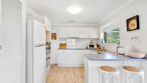A modern kitchen with white cabinetry, a central island with stools, a built-in oven, and a window with a view of greenery. - Beach Views - Spacious home by the ocean (Lennox Head)