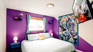Ein Schlafzimmer mit Bett, Nachttisch, Lampe, einem Fenster mit Vorhängen und einer Wand mit einem farbenfrohen Gemälde.