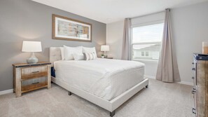 A bedroom with a large bed, two bedside tables with lamps, a framed artwork above the bed, a window with blinds, and a wooden dresser.