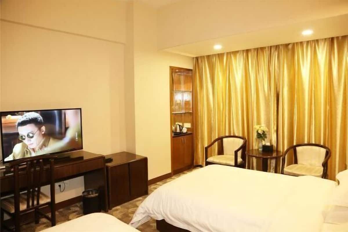 Een hotelkamer met een bed, een bureau met een televisie, een stoel, een tafeltje met een vaas en een raam met gordijnen.