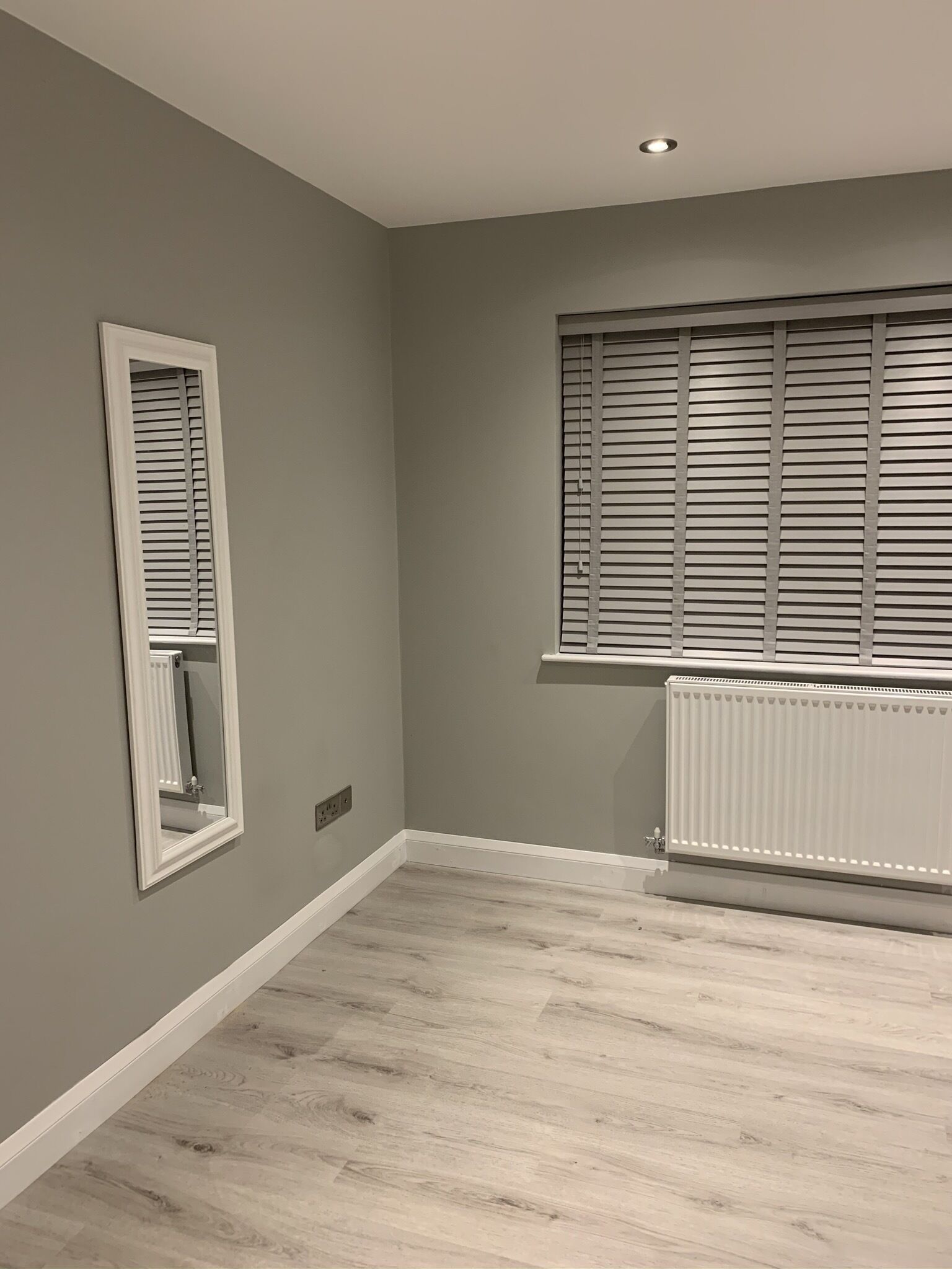 Une pièce avec un mur gris, une fenêtre avec des stores, un radiateur et un miroir accroché au mur.