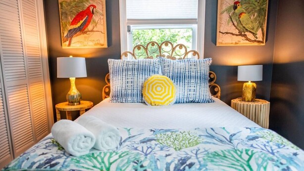 Una cama bien hecha con una colcha floral, dos almohadas y toallas enrolladas. Hay dos lámparas de pared a cada lado de la cama.