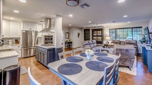Una cucina moderna con elettrodomestici integrati, un'area pranzo con tavolo in legno e sottopiatti blu, e un soggiorno accogliente con divano grigio e una grande finestra.