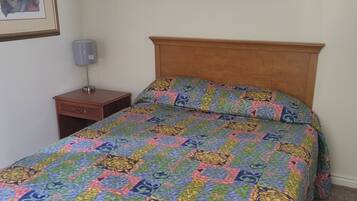 A bedroom with a wooden headboard, a colorful quilt, a small bedside table with a lamp, and a framed picture on the wall.