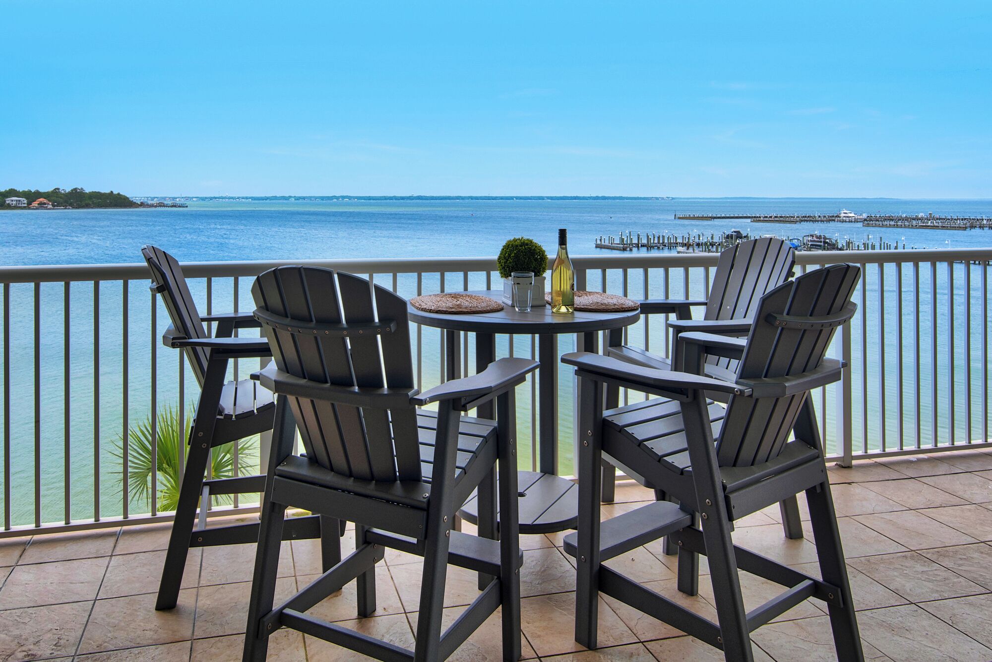 Ein Balkon mit vier Adirondack-Stühlen, einem runden Tisch und einer Weinflasche. Der Balkon bietet Blick auf ein Gewässer mit einem Pier im Hintergrund.