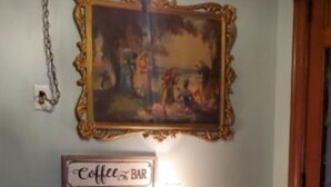 Een muur met een ingelijst schilderij, een bord waarop 'Coffee Bar' staat en een koffiezetapparaat op een tafel.