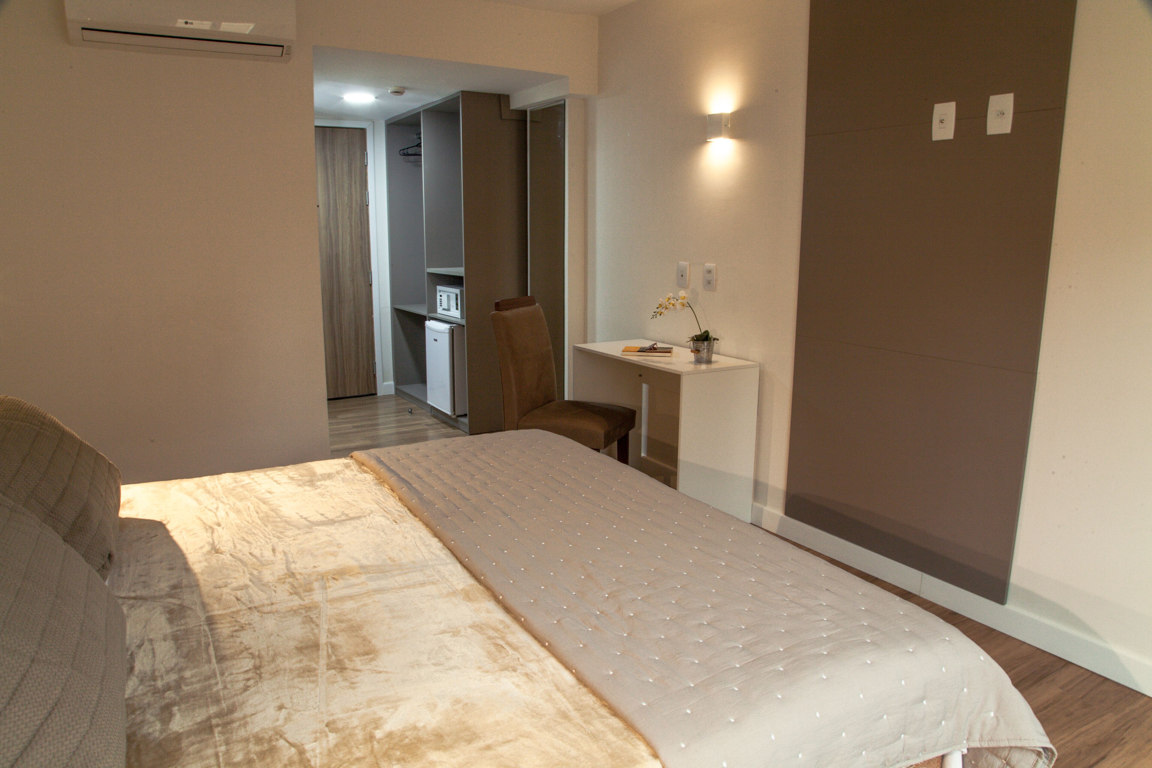 Un dormitorio con cama, escritorio y silla. Se ve una kitchenette al fondo.