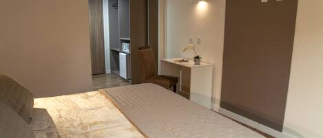 Un dormitorio con cama, escritorio y silla. Se ve una kitchenette al fondo.