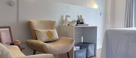 A modern living room with a beige armchair, a round wooden stool, a small table, and a white tablecloth-covered table.