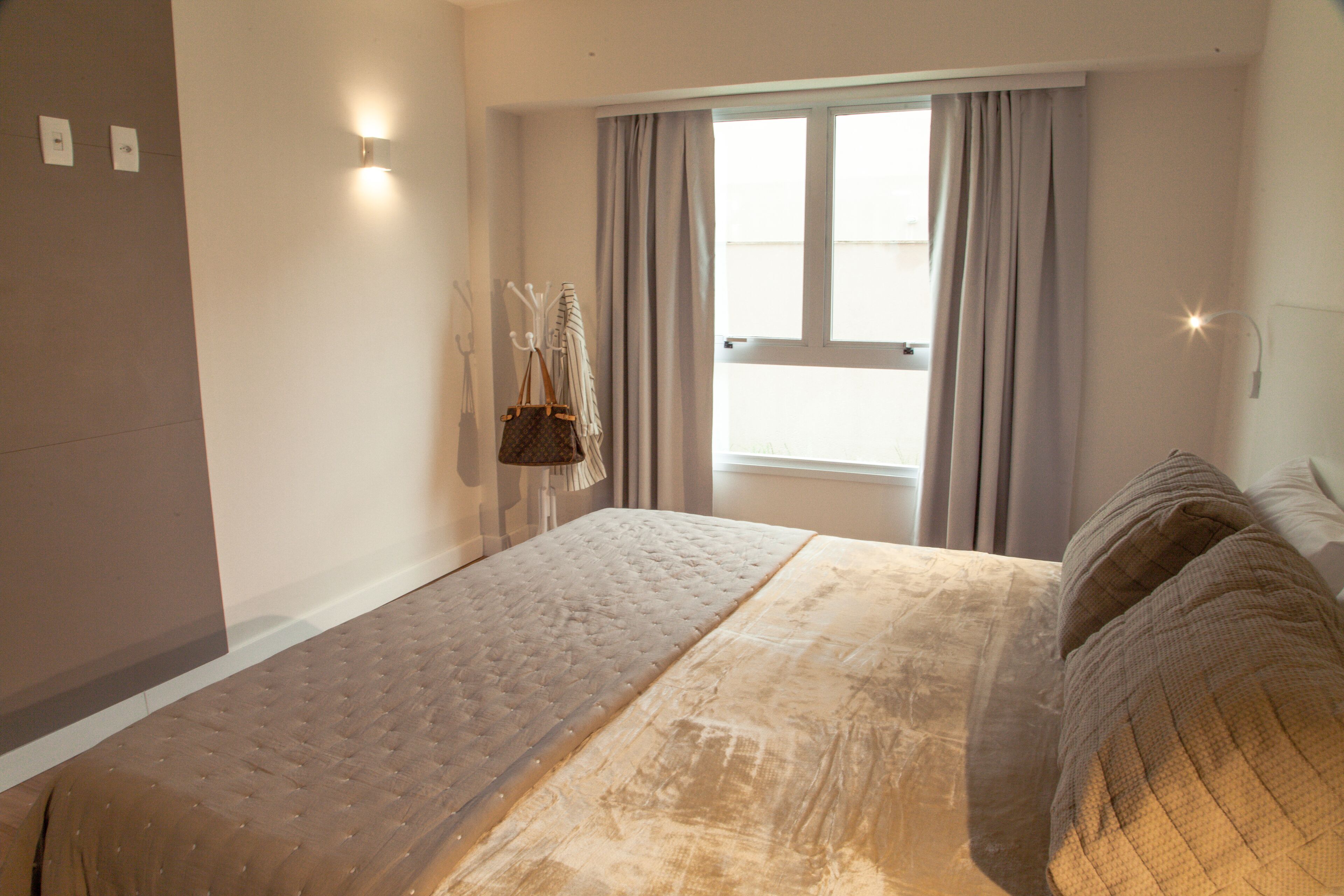 A hotel room with a bed, a window with curtains, a wall-mounted light, and a coat rack with a bag.