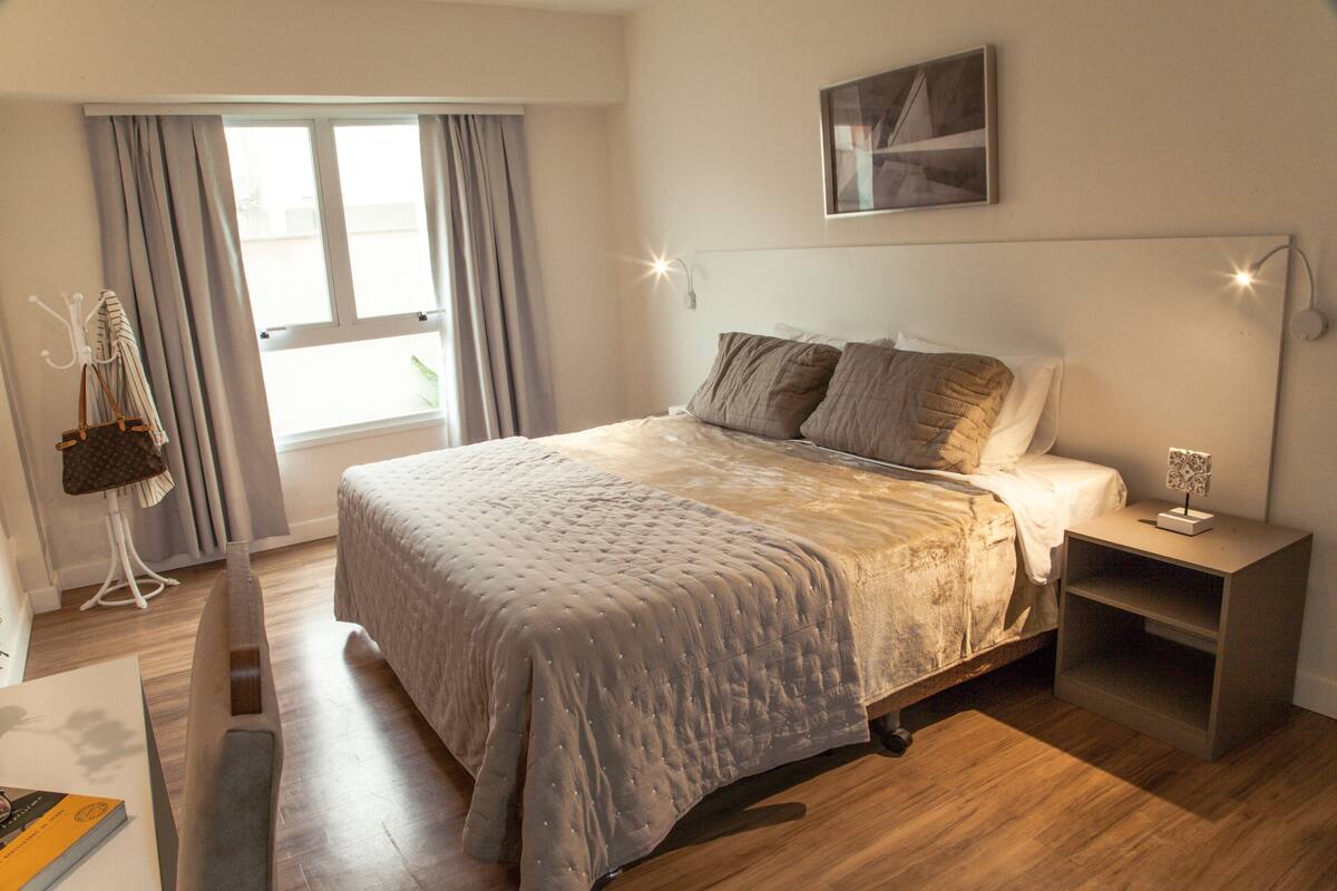 Een slaapkamer met een groot bed, een nachtkastje, een lamp en uitzicht op de omgeving door een raam.