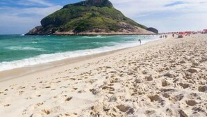 Une plage de sable parsemée d’empreintes, une grande colline verdoyante en arrière-plan et un ciel bleu clair.