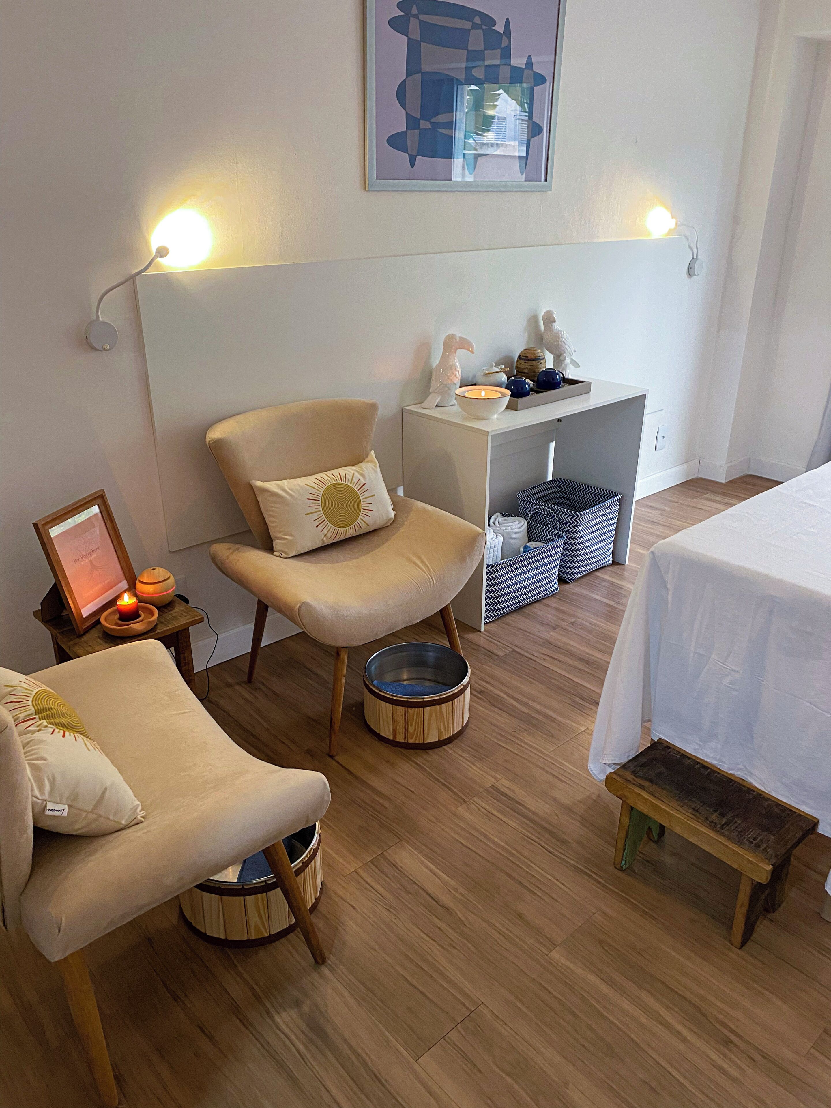 Una habitación acogedora con suelo de madera, un sillón color beige con un cojín con forma de sol, un taburete de madera pequeño y una cama blanca con colcha blanca.