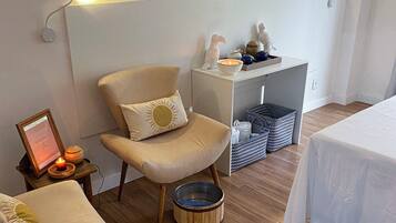 Una habitación acogedora con suelo de madera, un sillón color beige con un cojín con forma de sol, un taburete de madera pequeño y una cama blanca con colcha blanca.