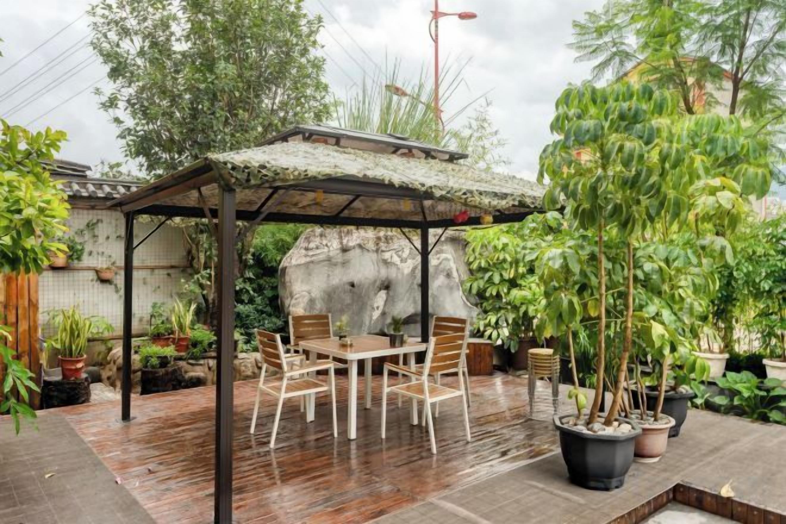 Et overdækket udendørs siddeområde med et bord og stole, omgivet af plantekasser og en klippeformation.