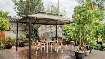 Et overdækket udendørs siddeområde med et bord og stole, omgivet af plantekasser og en klippeformation.
