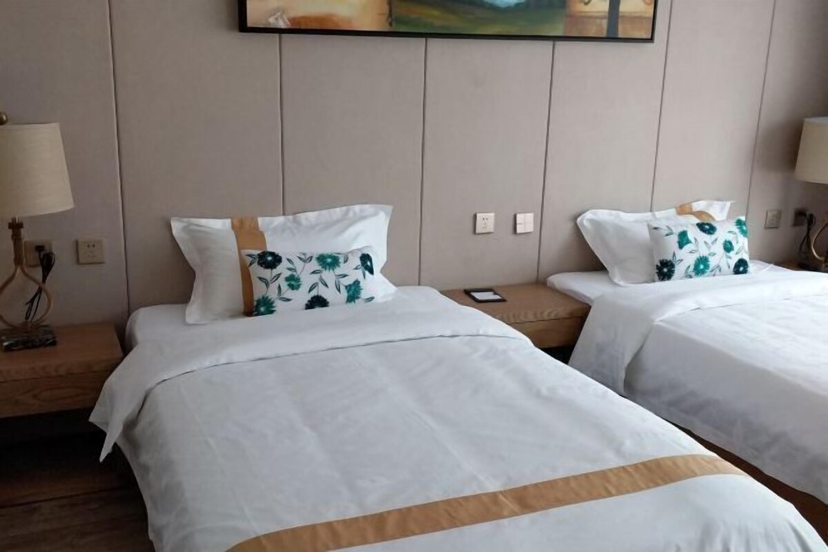 A hotel room with two beds, each with white bedding and floral-patterned pillows. There is a wooden nightstand with a lamp on the left side.