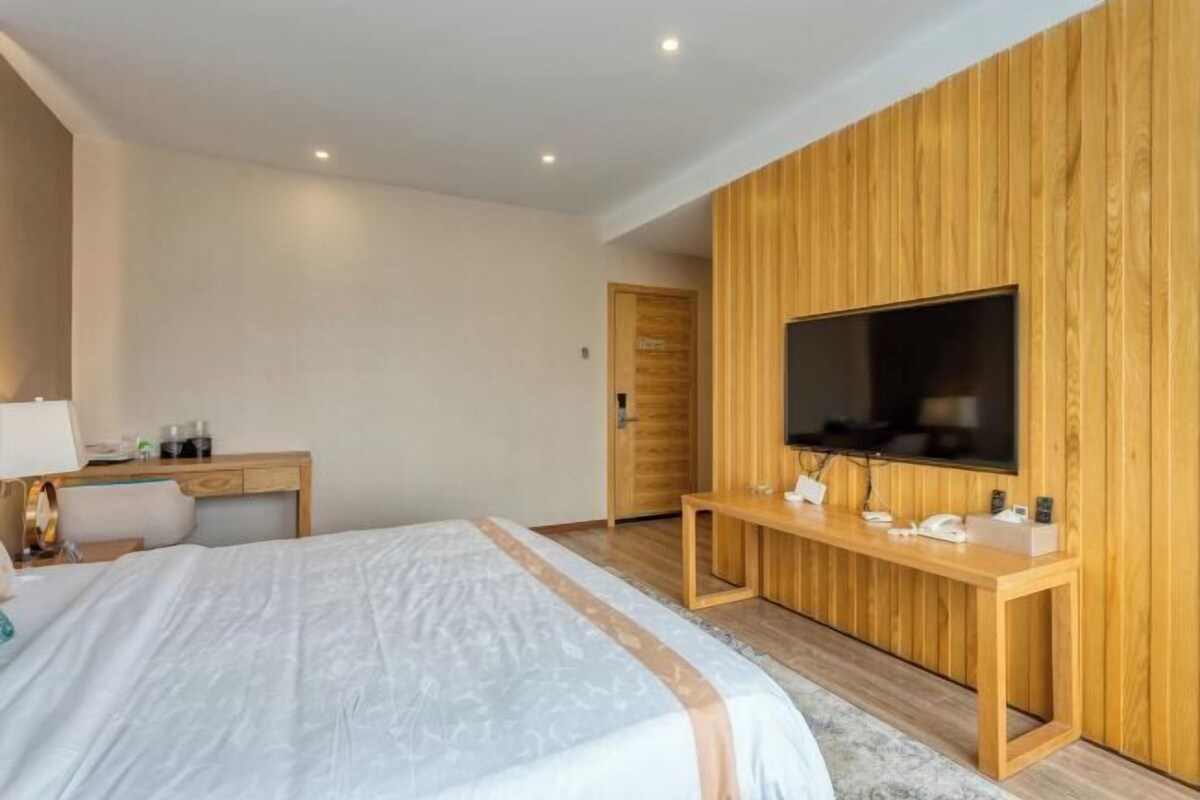 A modern hotel room with a large bed, a wooden desk, a flat-screen TV mounted on the wall, and a wooden wardrobe.