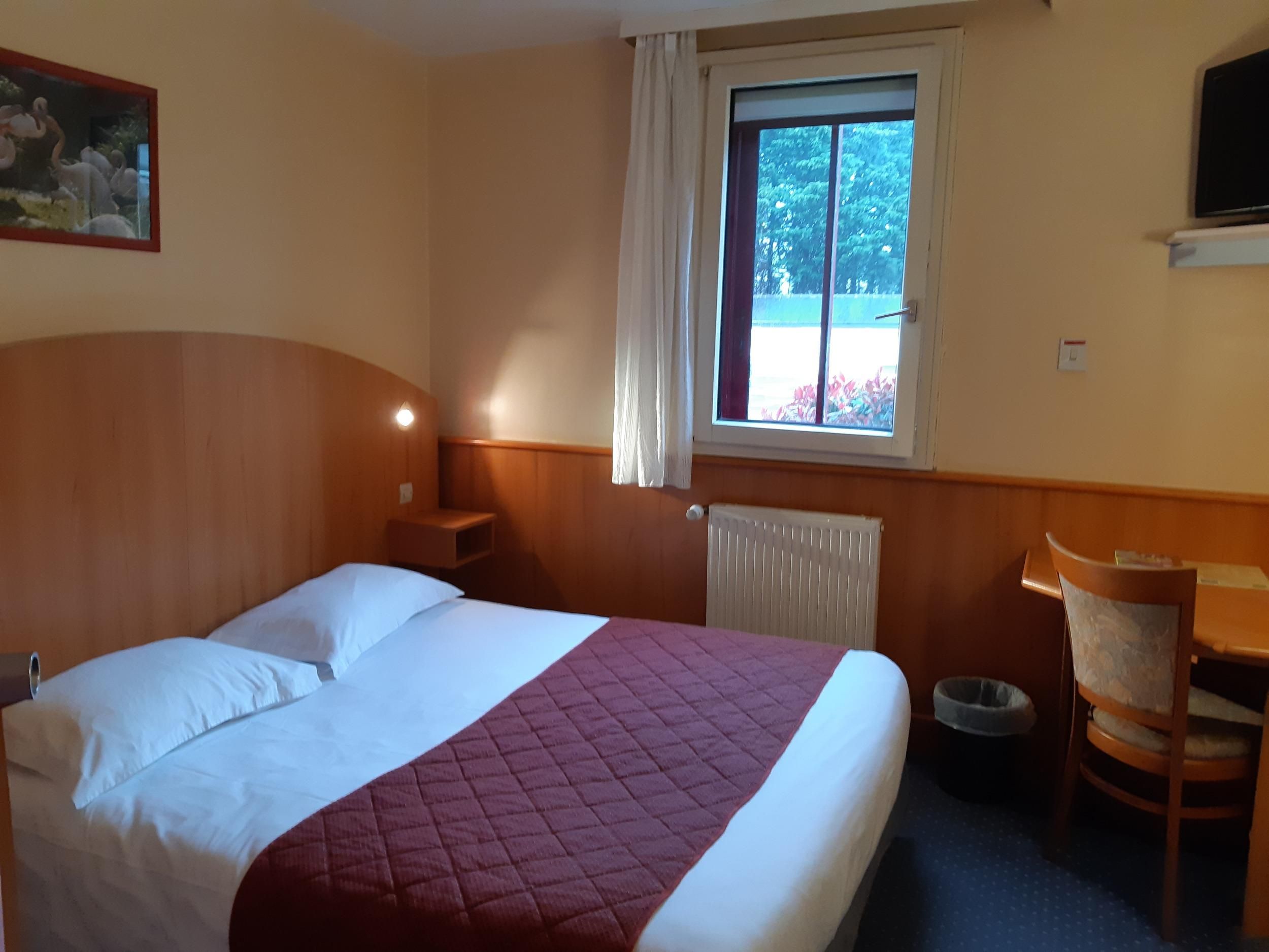 Habitación de hotel con cama, escritorio, silla, ventana y vista a vegetación, además de un radiador.