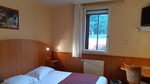 A hotel room with a bed, a desk, a chair, a window with a view of greenery, and a radiator. - Logis Hotel Lucotel (Lanvollon)