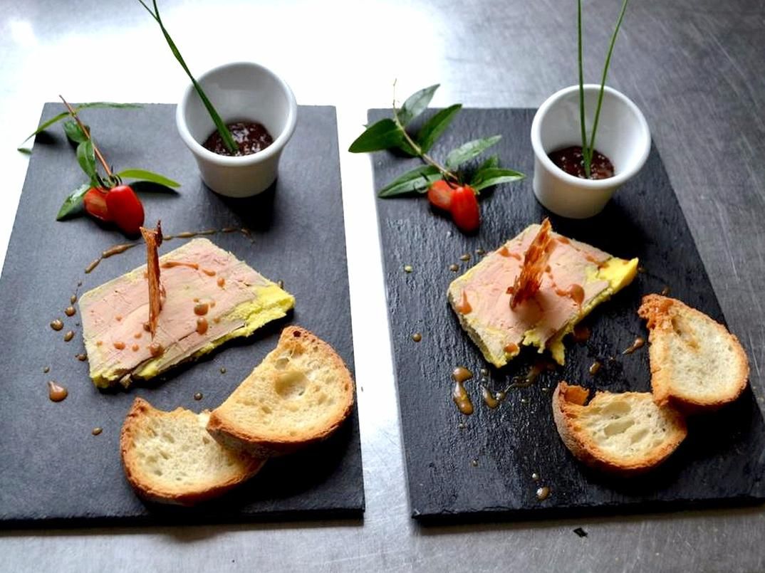 Two square black slate boards with food items and bread, each accompanied by a small white bowl with a plant.