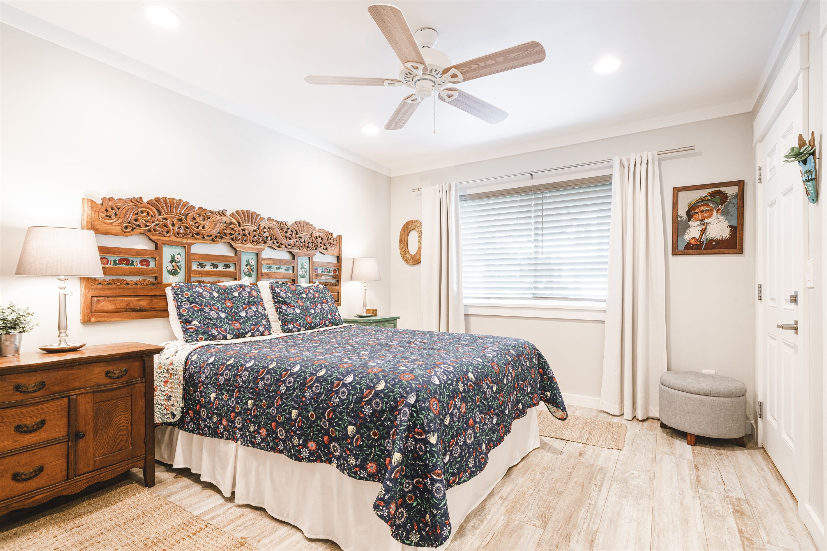 Une chambre à coucher comprenant un lit, une tête de lit en bois, un ventilateur de plafond, une table de chevet avec une lampe et une fenêtre avec des rideaux.