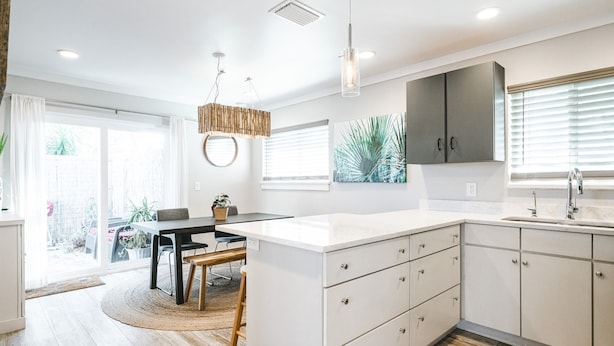 Una cocina moderna con armarios blancos, una zona de comedor con una mesa redonda y un amplio ventanal con persianas.
