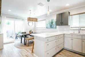 Una cocina moderna con armarios blancos, una zona de comedor con una mesa redonda y un amplio ventanal con persianas.