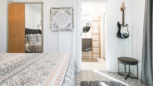 Un dormitorio con una colcha estampada, un espejo, una guitarra y un taburete pequeño.