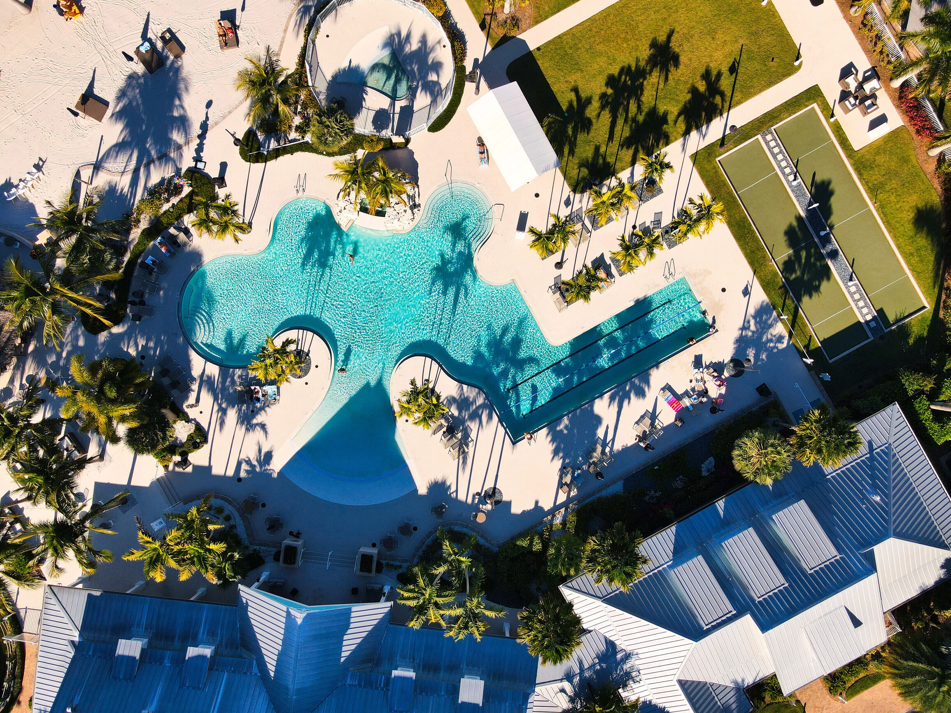 Vue aérienne d’un complexe hôtelier doté d’une grande piscine en forme de guitare, entourée de palmiers et de chaises longues.