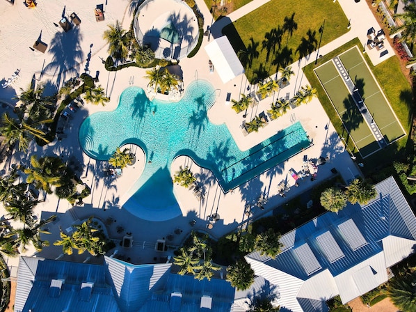 An aerial view of a resort with a large pool shaped like a guitar, surrounded by palm trees and sun loungers.