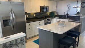 A modern kitchen with a stainless steel refrigerator, white cabinets, a black backsplash, and a large island with bar stools. - Modern large 2 bedroom/2 bath Condo on Intercoastal with gorgeous  sunset views (St Pete Beach)