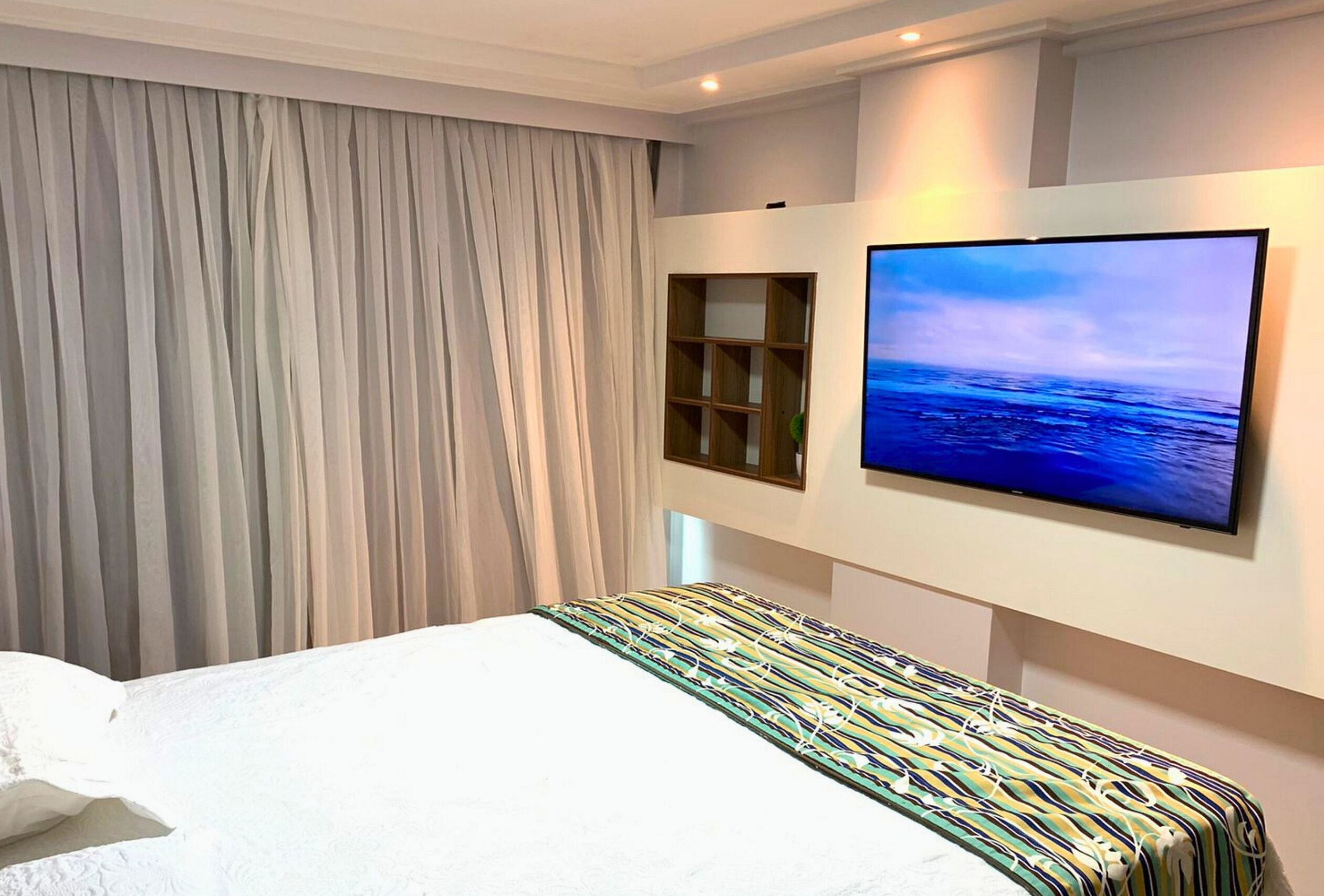Een hotelkamer met een bed, een groot raam met gordijnen, een flatscreen televisie aan de muur en een klein houten plankje.