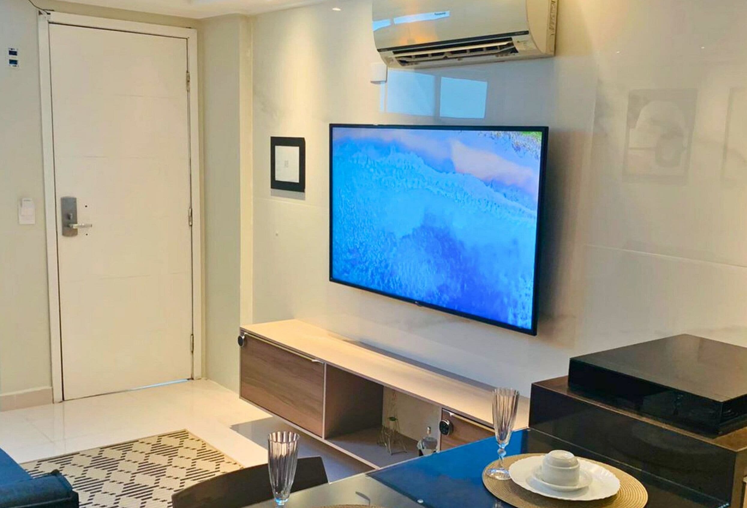 Een moderne woonkamer met een flatscreen televisie aan de muur, een houten console en een vloerkleed met dessin, plus een glazen vaas.