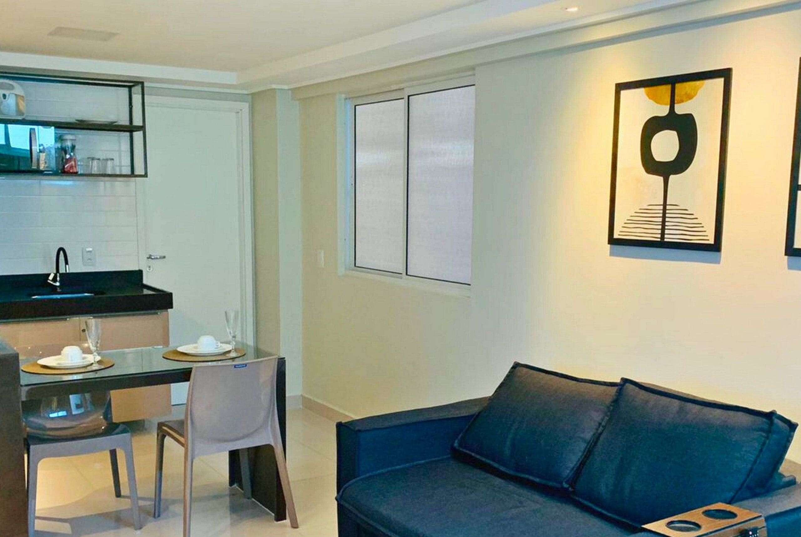 A modern living room with a dark blue sofa, a dining area with a table and chairs, and a kitchen area with a black countertop and white cabinets.