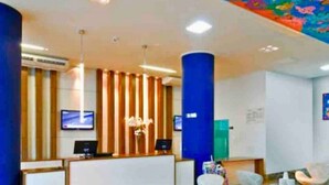 Un área de recepción moderna con un mostrador de madera, dos monitores y un jarrón decorativo. Hay varias sillas blancas y un pilar azul.