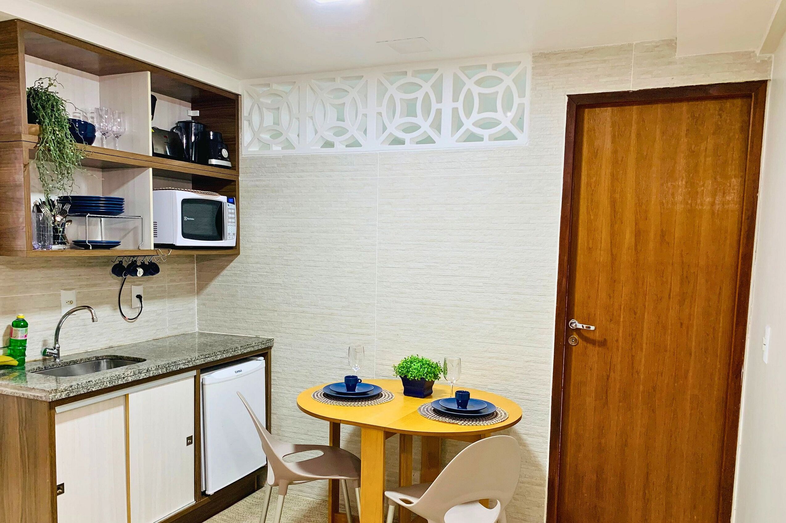 A compact kitchen with a microwave, sink, and cabinets. There is a dining table with chairs and a small potted plant.