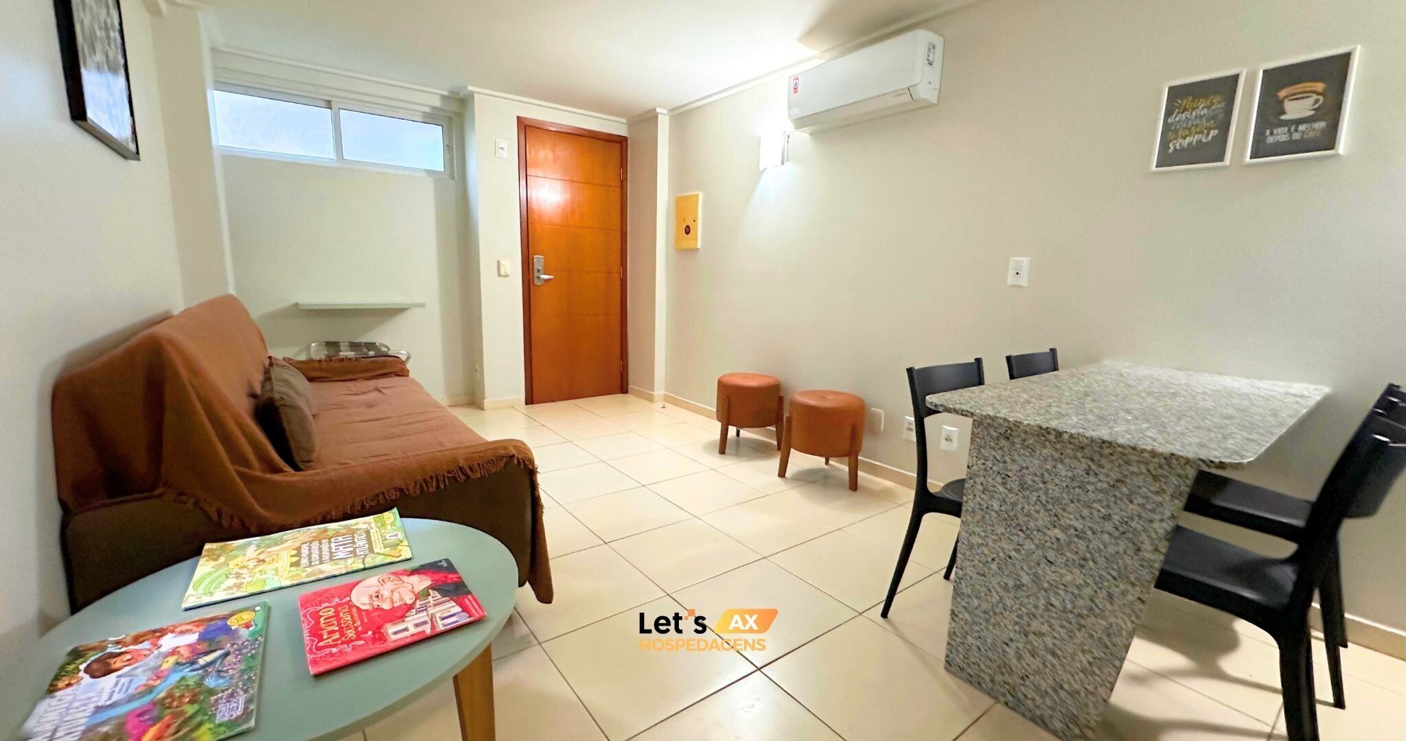 A small, well-lit room with a sofa, a dining table with chairs, and a small kitchen area. There is a window providing natural light and a door leading to another room.